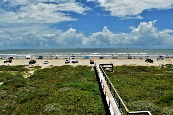 A scenic beach view with parked cars, a wooden walkway, greenery, and a cloudy sky above the ocean waves.