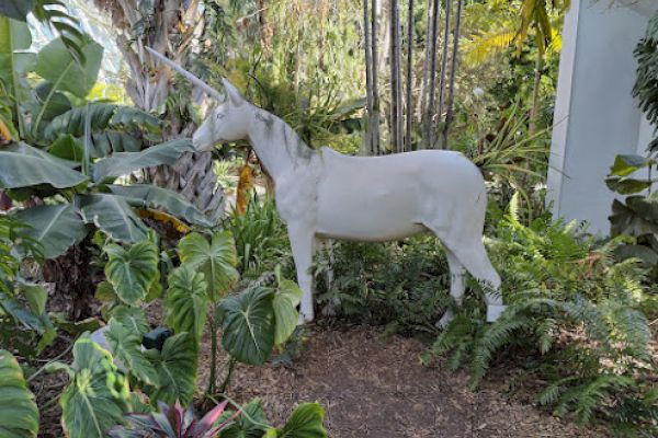 A white unicorn statue is surrounded by lush green plants and trees in a garden setting.