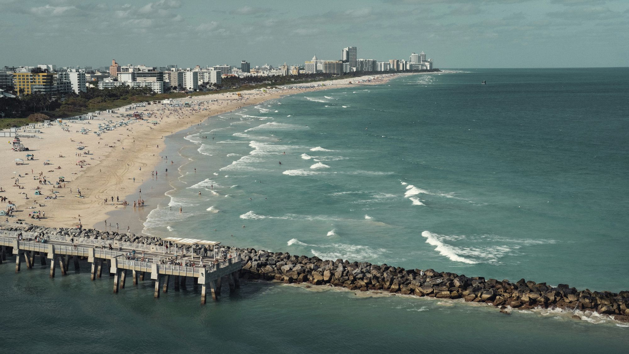 A beach with a pier and people, bordering a city skyline; waves crash against the shore under a partly cloudy sky.