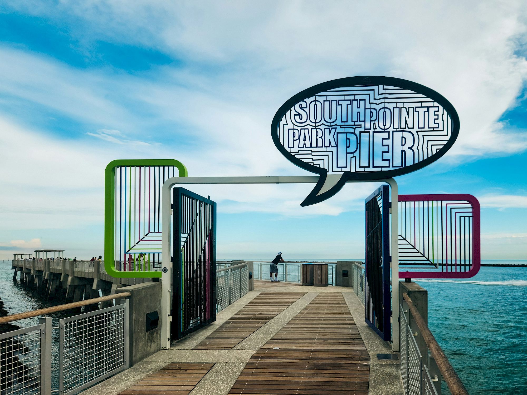 A pier with a sign reading "South Pointe Park Pier," surrounded by the ocean and a partly cloudy sky.