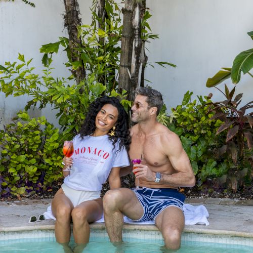Two people are sitting by a pool, holding drinks, surrounded by greenery and plants.