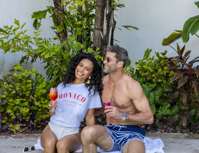 Two people are sitting by a pool, holding drinks, surrounded by greenery and plants.